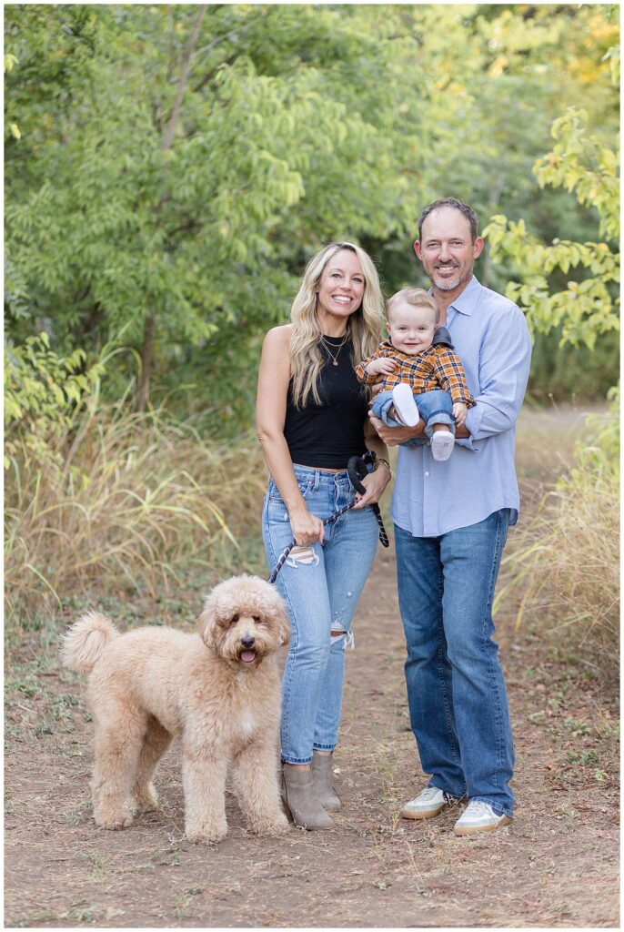Texas family photographer, Wisp + Willow Photography Co., captures fall mini sessions at Frisco Commons.  A father holds his baby son who is smiling at the camera sitting up in his Dad's arms.  His Mom stands right next to him who wears a black tank top and jeans and holds their doodle dog on a black leash.